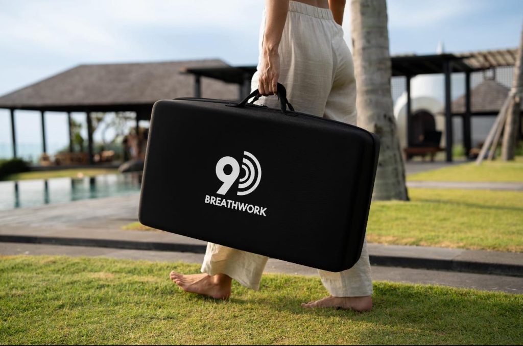 A barefoot person in light pants carries a black case labeled 9 BREATHWORK by a poolside, with palm trees and a modern outdoor building in the background.
