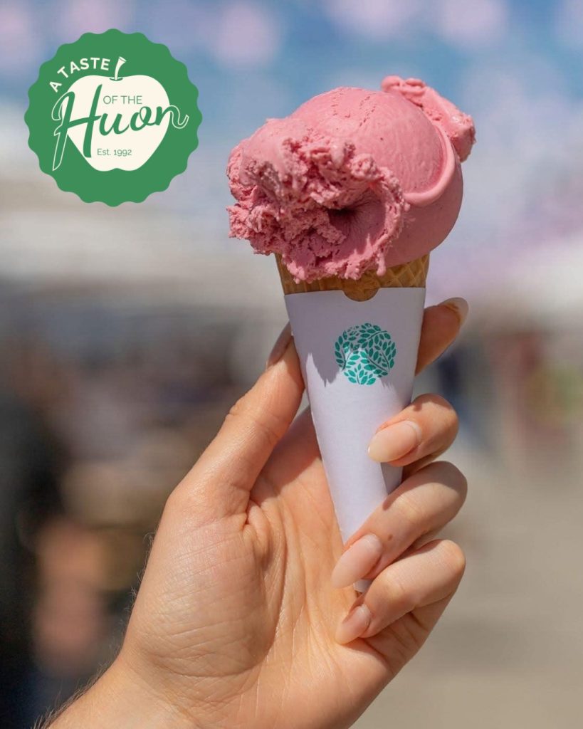 A hand holding a waffle cone with a scoop of pink ice cream, wrapped in a white paper sleeve. The background is blurred, and a green A Taste of the Huon festival logo is in the top left corner.