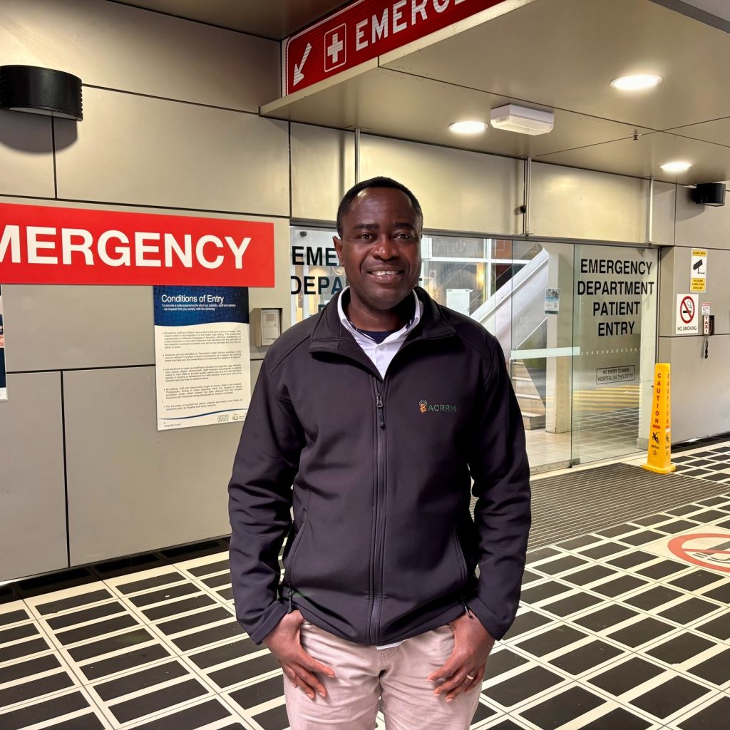 A man in a black jacket stands and smiles in front of the entrance to a hospital emergency department. Red and white emergency signs and glass doors are visible in the background.