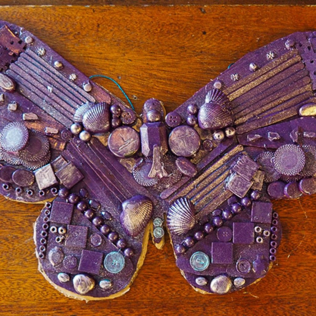 A butterfly-shaped collage made from assorted small objects, painted purple, including beads, buttons, shells, and textured pieces, displayed on a wooden surface.