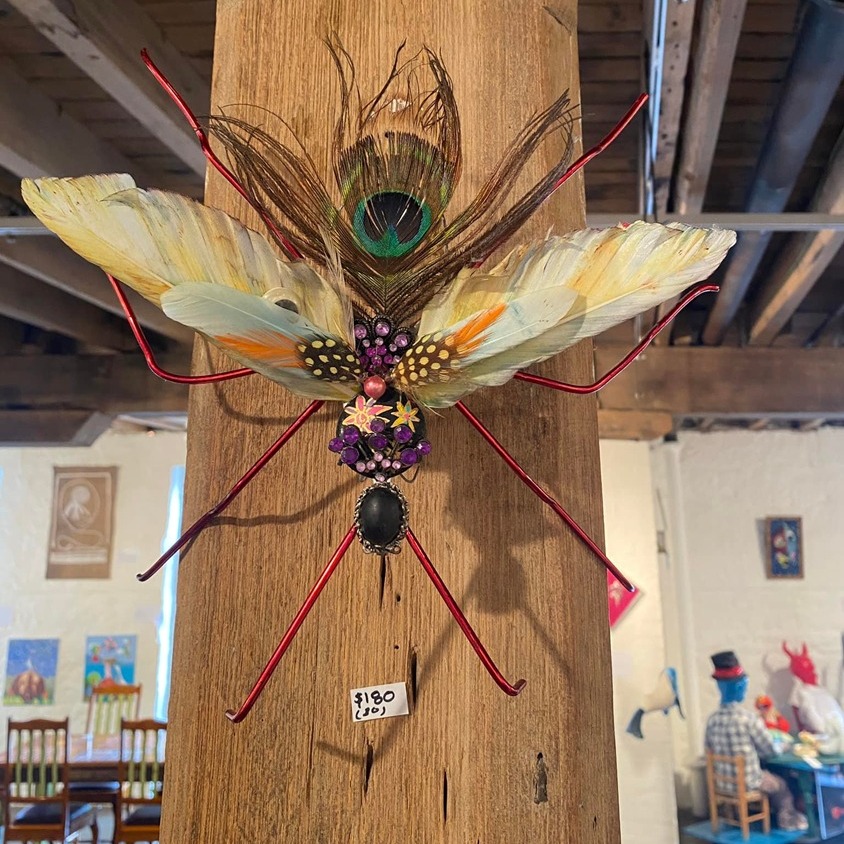 A decorative, colorful insect sculpture with feather wings and wire legs is mounted on a wooden post indoors. It has a peacock feather on top and costs $180. Blurred art and people are visible in the background.
