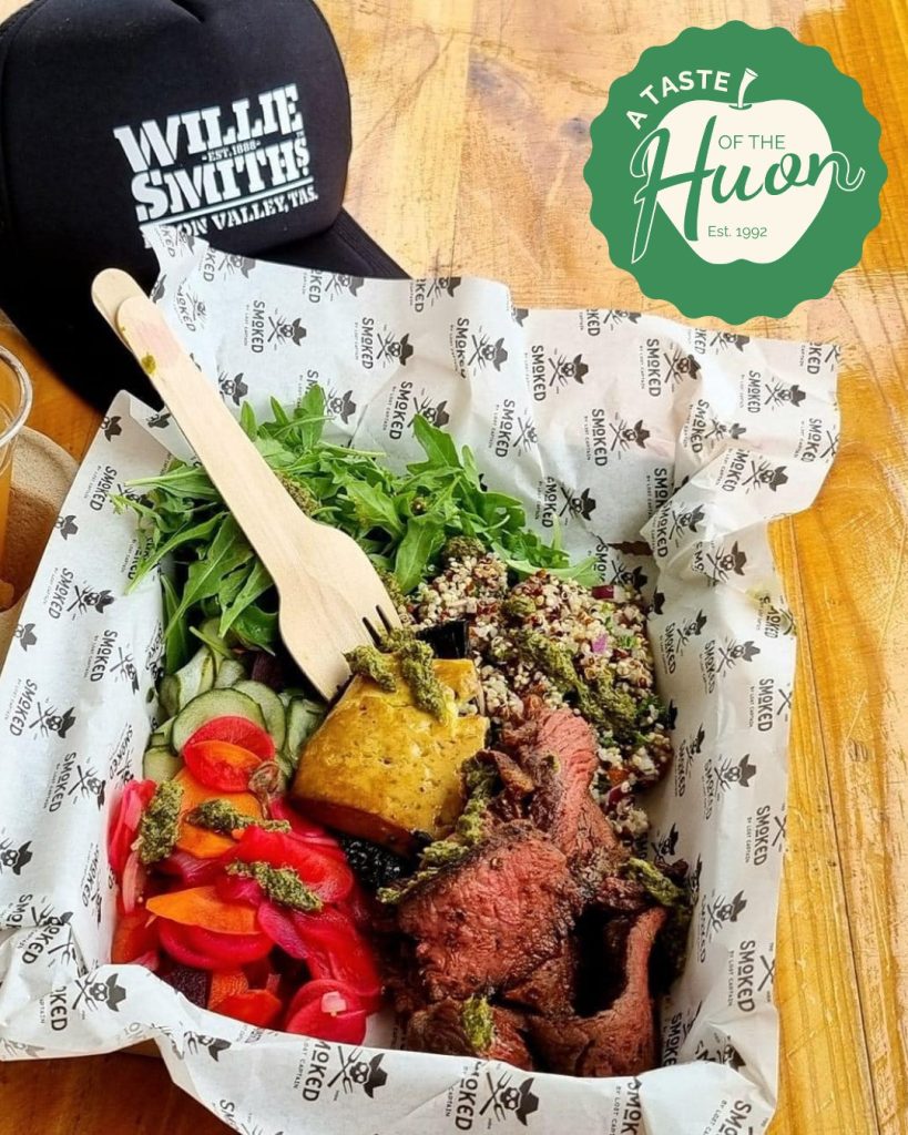 A tray with sliced steak, roasted potato, mixed greens, quinoa salad, and roasted red peppers, served with a wooden fork. A black Willie Smiths cap and A Taste of the Huon logo are visible in the background.