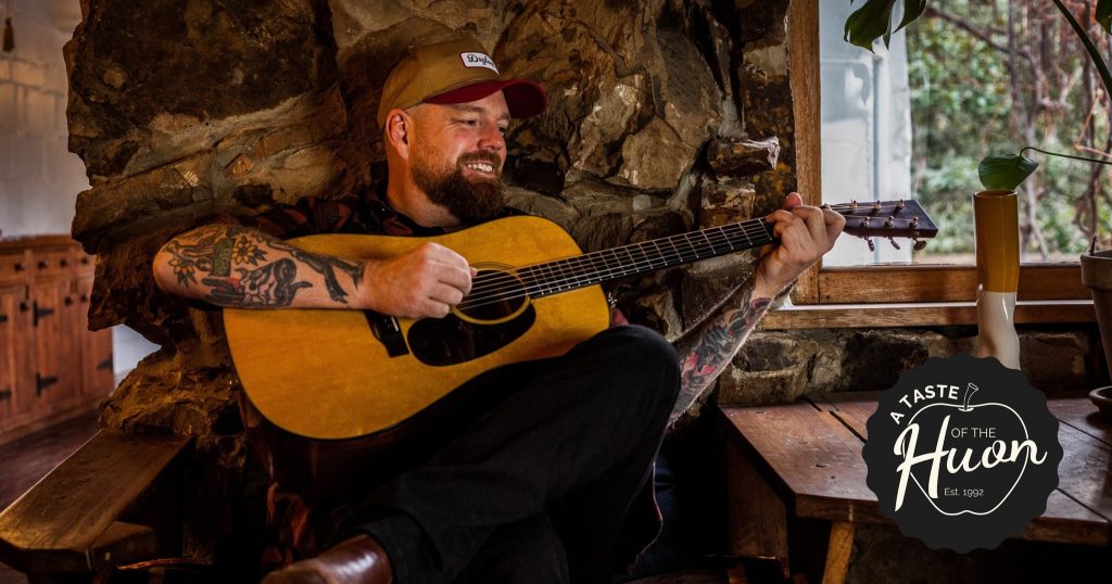 A bearded man with tattoos plays an acoustic guitar while smiling and sitting in a rustic room with a stone fireplace. A circular “A Taste of the Huon” logo is in the bottom right corner.