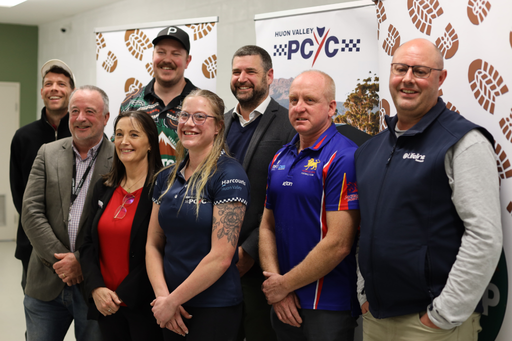 Eight smiling adults stand together indoors in front of banners for Huon Valley PCYC. The group includes men and women dressed in casual and business attire, and they appear to be at an event or community gathering.