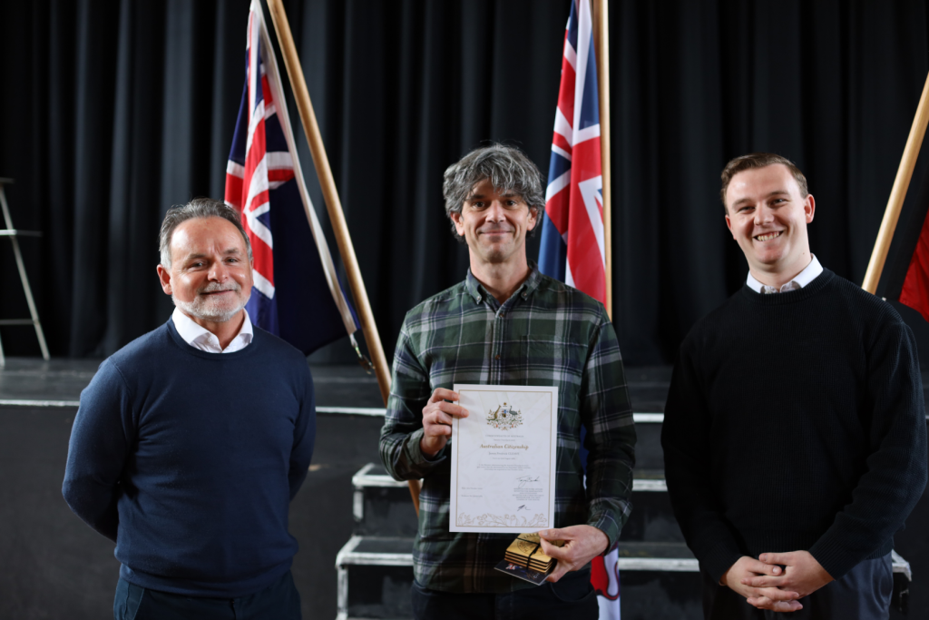 Three men stand indoors in front of British flags. The man in the middle holds a certificate and small box, smiling, while the other two men stand on either side, also smiling. A black curtain and steps are in the background.