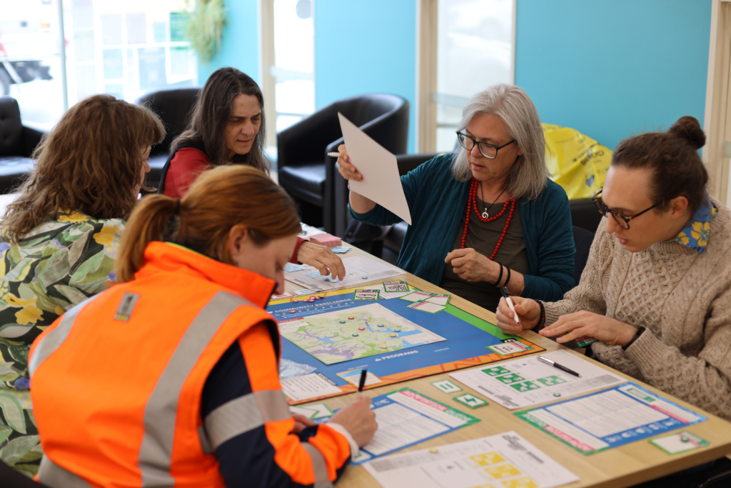 Five women sit around a table engaged in an activity with papers, pens, and a colorful board game or planning materials. One woman in an orange vest writes while another holds up a sheet of paper.