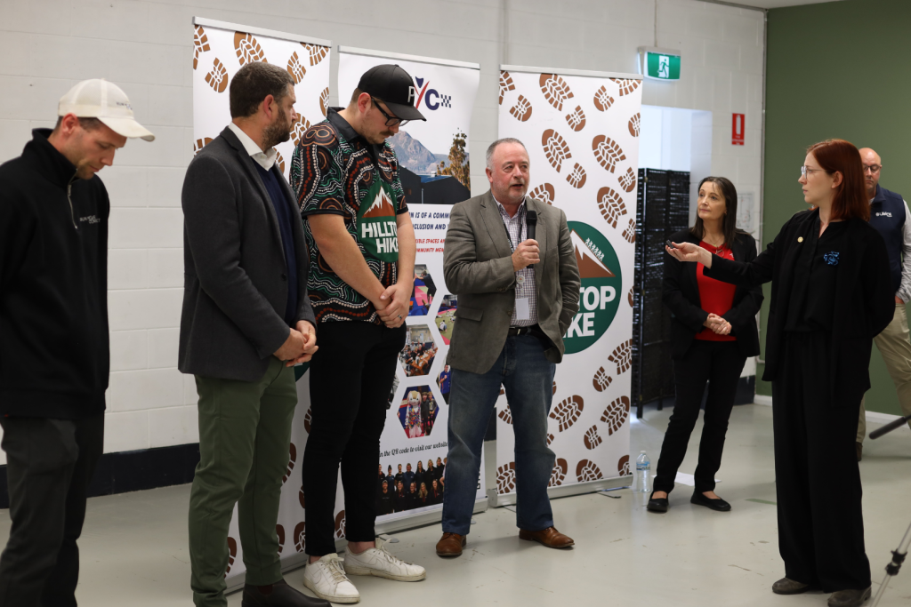 A group of six people stand in front of banners indoors. One man holds a microphone and speaks while others listen. The group is dressed in a mix of business and casual attire.