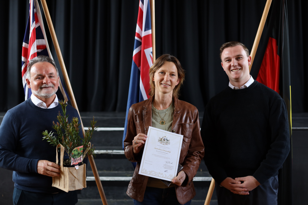 Three people stand indoors in front of flags. The woman in the center holds a certificate, the man on the left holds a native plant in a basket, and the man on the right stands with his hands clasped, all smiling at the camera.