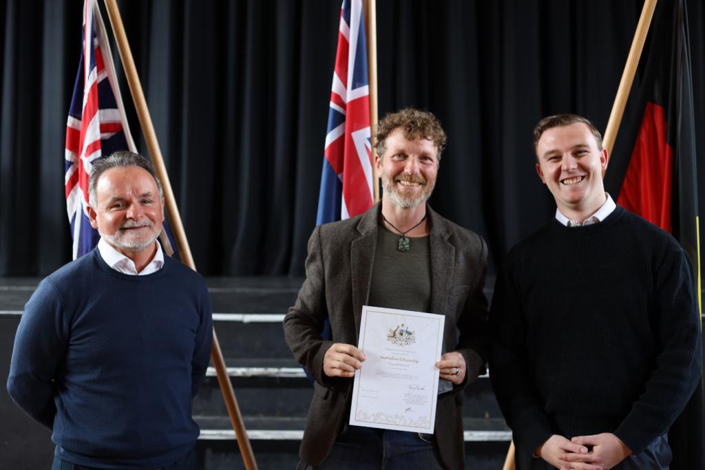 Three men stand smiling indoors in front of steps and flags. The man in the center holds an Australian citizenship certificate. The background features a dark curtain and both Australian and Aboriginal flags on display.