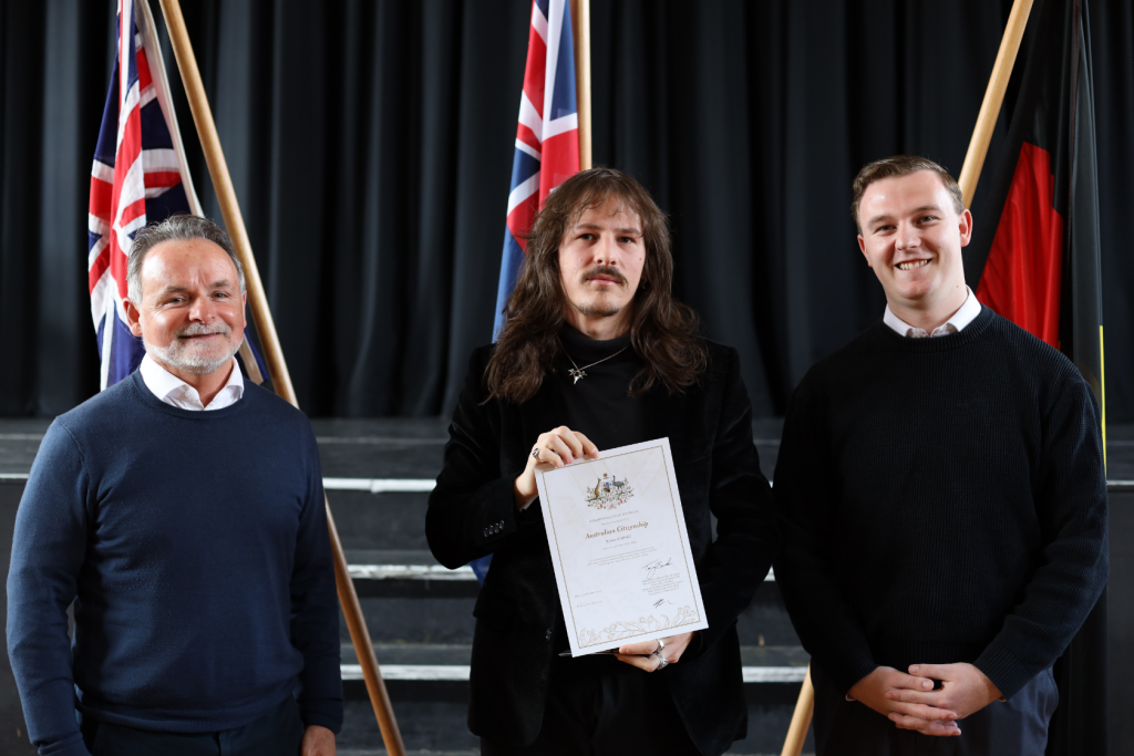 Three people posing indoors in front of Australian and Aboriginal flags. The person in the center holds a certificate and stands between two smiling people. They are on a stage with black curtains and steps behind them.