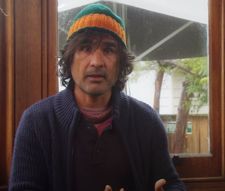 A man with wavy hair wearing a colorful knit beanie, a maroon shirt, and a dark blue sweater sits indoors by a window, looking slightly concerned while gesturing with his hands.