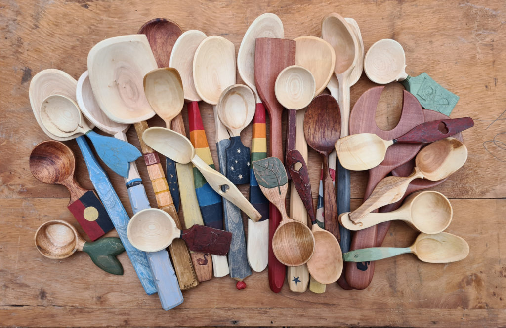 A collection of handmade wooden spoons and utensils in various shapes, sizes, and colors, arranged on a wooden surface. Some handles are painted or carved with unique designs.