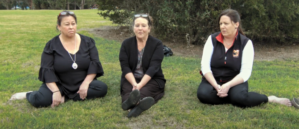Three women are sitting cross-legged on grass in a park, facing the camera. They appear relaxed and are dressed in casual clothing, with trees and bushes in the background.