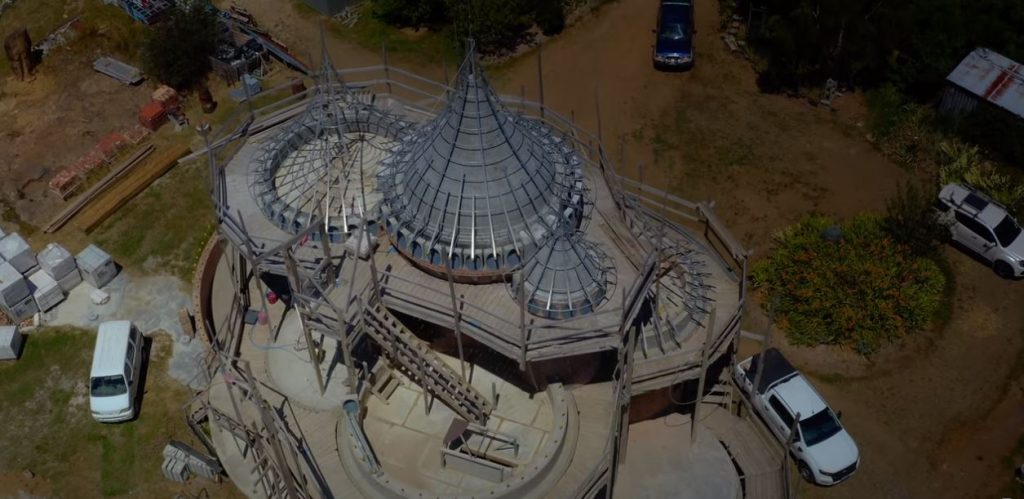 Aerial view of a unique round building under construction, featuring conical metal frameworks for roofs, surrounded by dirt paths, parked vehicles, and scattered construction materials.