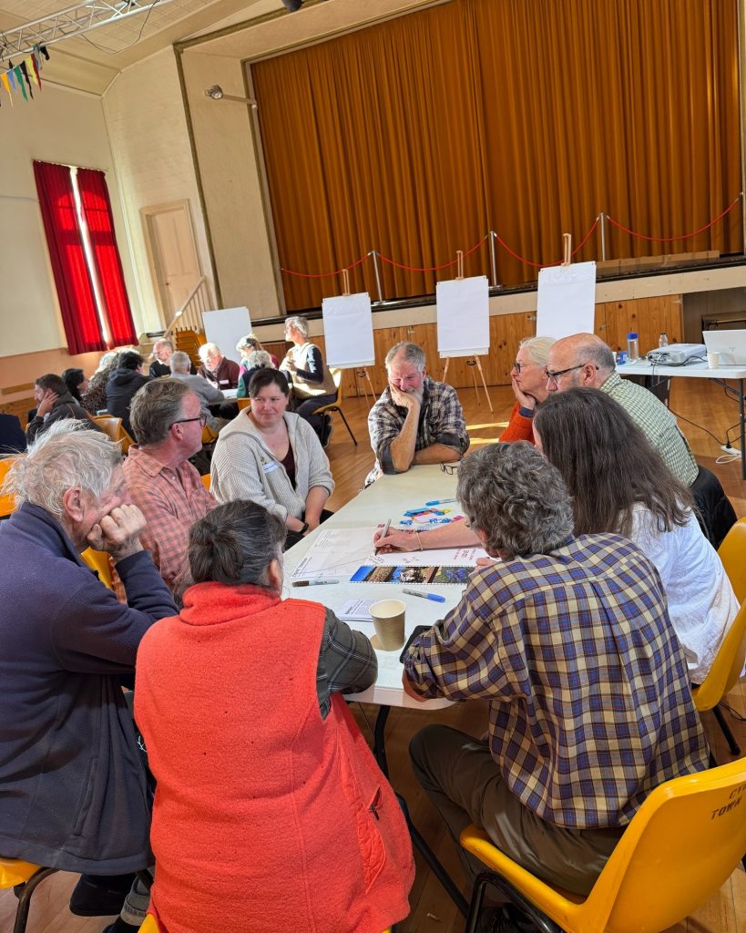 A group of people sit around a table in a brightly lit hall, engaged in discussion. Flipcharts and markers are on the table, and blank easels stand near a stage with orange curtains in the background.