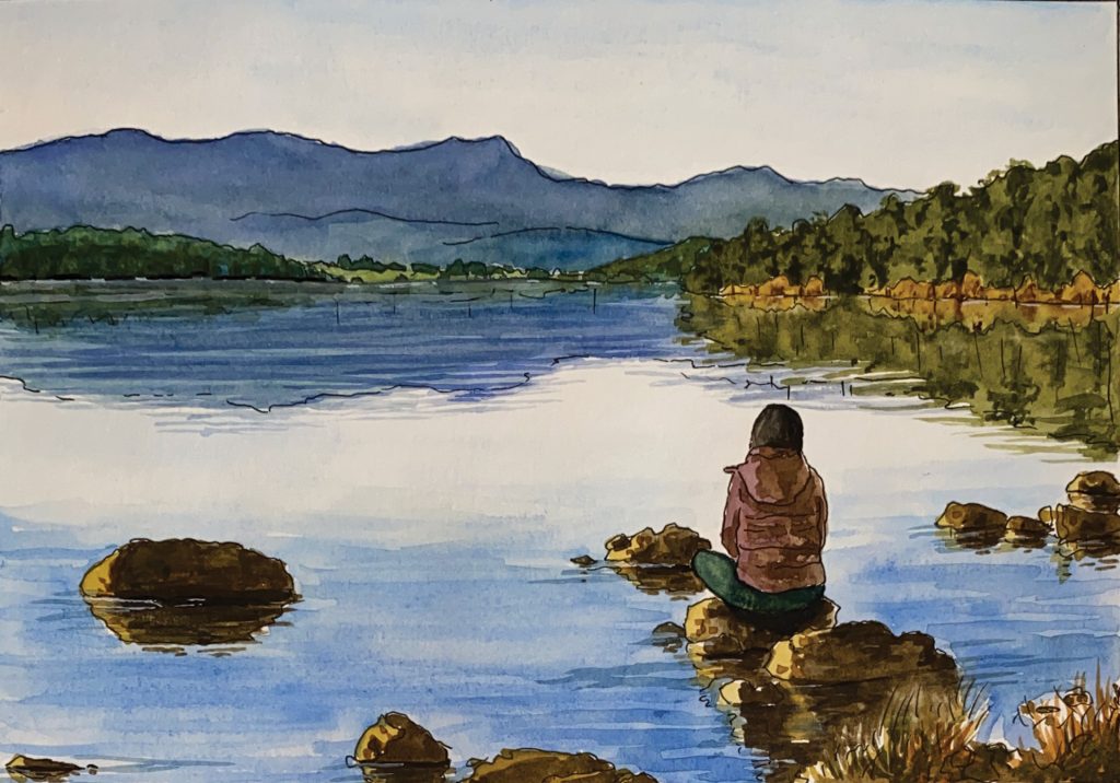A person sits alone on rocks at the edge of a calm lake, facing distant mountains under a blue sky. Trees line the shore, and the water reflects the landscape quietly.