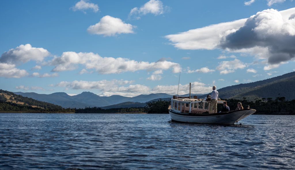 A small boat with people on board floats on a calm lake, surrounded by forested hills and distant mountains under a partly cloudy blue sky.