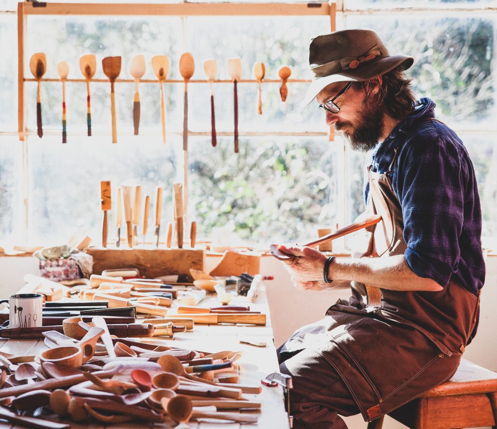 A bearded person wearing glasses, a hat, and an apron carves a wooden spoon at a sunlit workbench filled with handmade wooden utensils, with more utensils hanging on the wall behind.