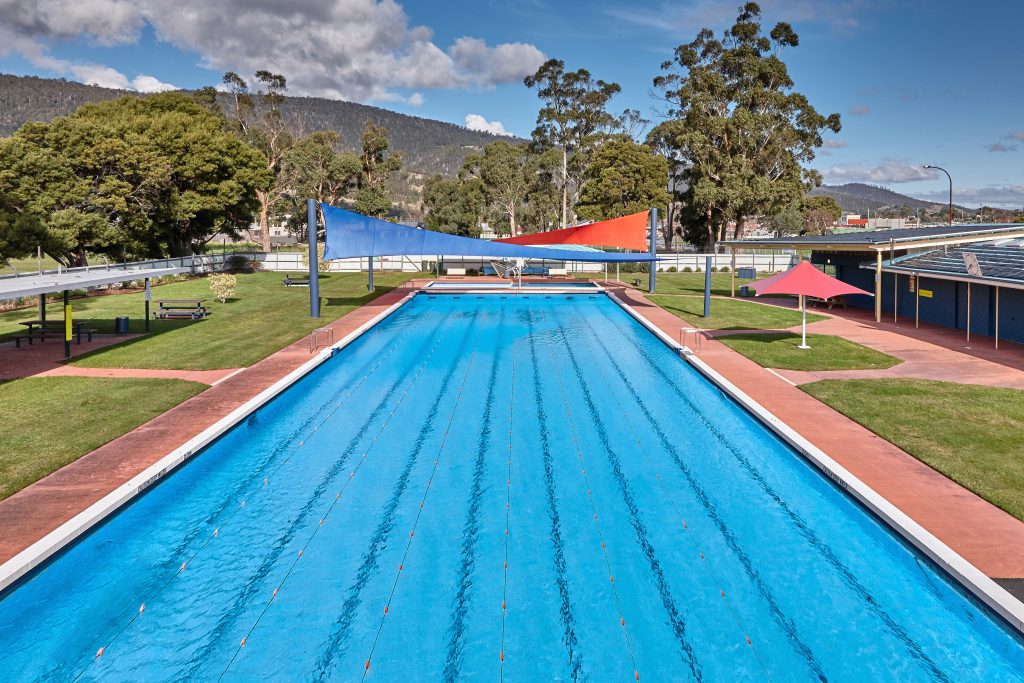 An outdoor swimming pool with clear blue water, lane markers, sunshades above, and green grass surrounding the pool. Trees, a bench, and distant hills are visible under a partly cloudy sky.