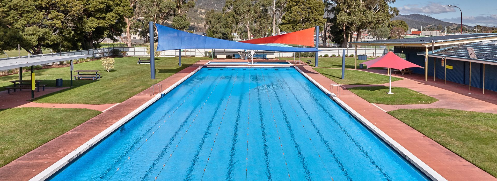 An outdoor swimming pool with clear blue water, lane markers, sunshades above, and green grass surrounding the pool. Trees, a bench, and distant hills are visible under a partly cloudy sky.