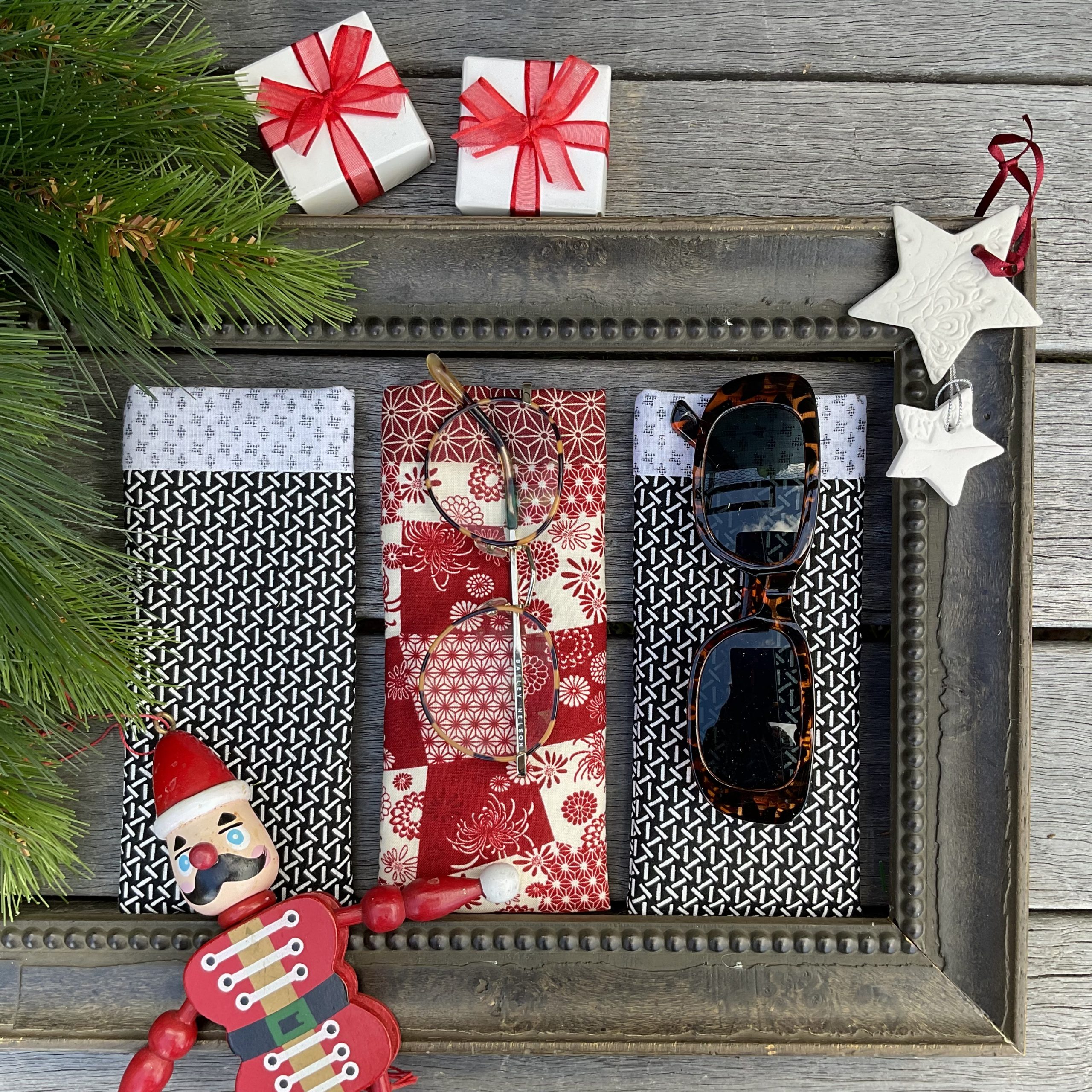 A festive flat lay with glasses cases in a wooden frame, a nutcracker ornament, sunglasses, white star ornaments, pine branches, and two small gift boxes with red ribbons on a wooden surface.