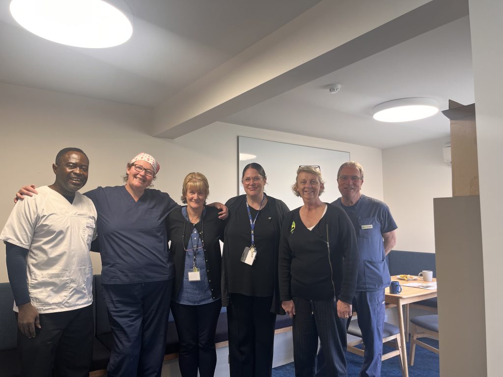 Six smiling healthcare workers, some in scrubs and others in casual or work attire, stand together in a well-lit room with a table and chairs in the background.