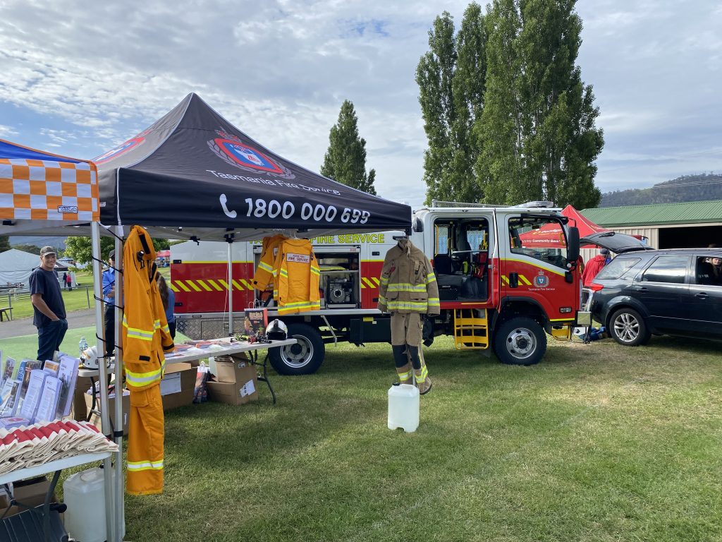 A fire service display at an outdoor event features a fire truck, a canopy tent with fire service branding, firefighting gear, informational materials, and a person in firefighting gear standing nearby on grass.