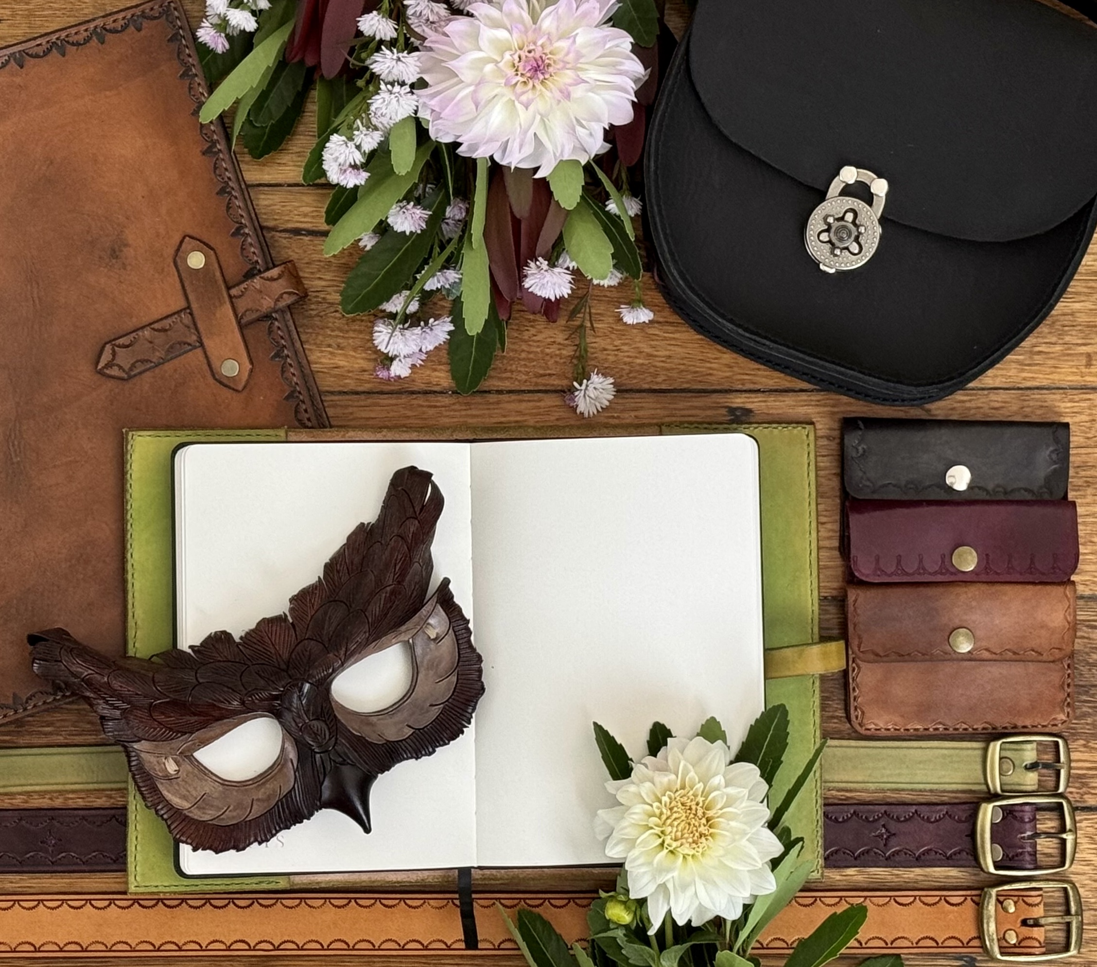 A flat lay of leather goods, an open blank journal with an owl mask on top, flowers, belts, a black satchel, and small leather pouches, all arranged on a wooden surface.