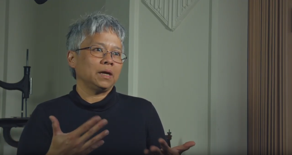 A person with short gray hair and glasses, wearing a dark top, gestures with their hands while speaking in an indoor setting with neutral-colored walls.