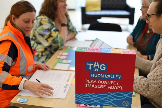 Four people sit around a table, discussing papers and materials. In the foreground, a colorful box labeled TAG HUON VALLEY: PLAY. CONNECT. IMAGINE. ADAPT. is prominently displayed.