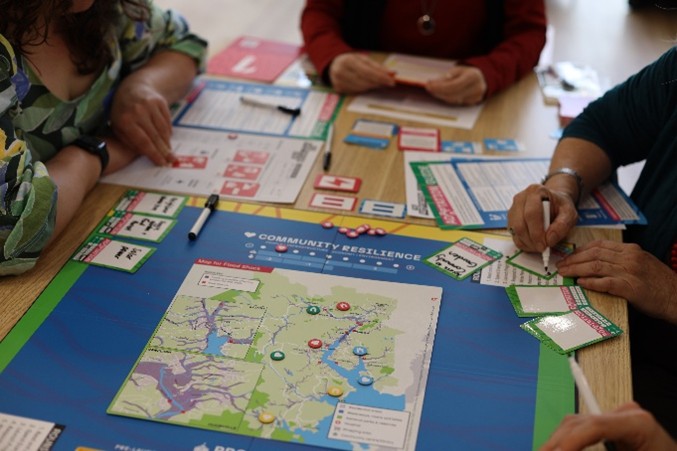 Three people are playing a tabletop board game called Community Resilience, which features a colorful map, game cards, tokens, and player sheets on a wooden table. Hands are visible as players mark information and move pieces.