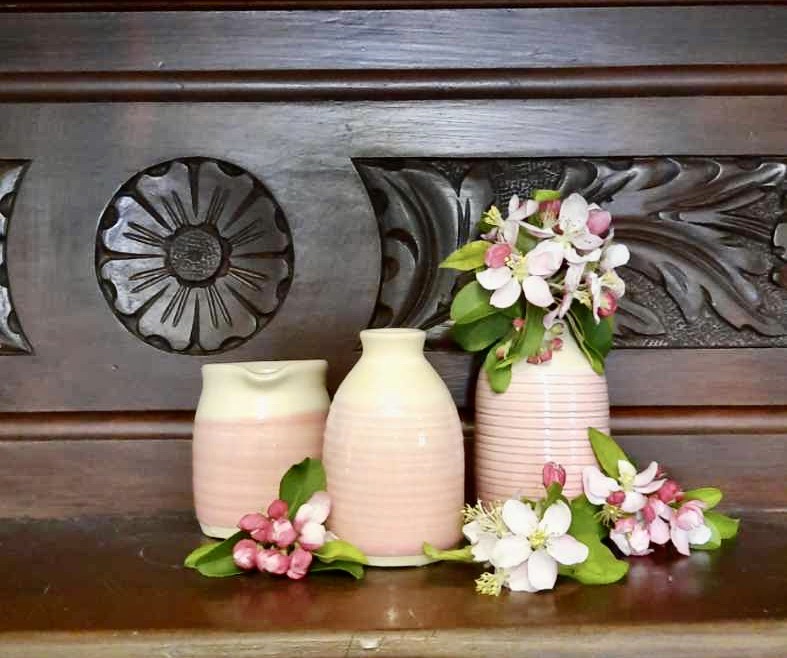 Three pink and cream ceramic vases, two small and one tall, are arranged on a dark carved wooden shelf, with pink and white blossoms and green leaves tucked among them.