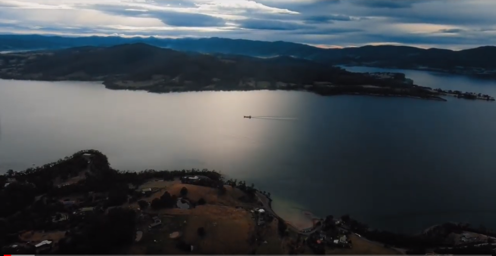Aerial view of a lake at dusk with a small boat leaving a wake across the water, surrounded by hills and scattered houses, under a dramatic cloudy sky with sunlight reflecting off the lake.