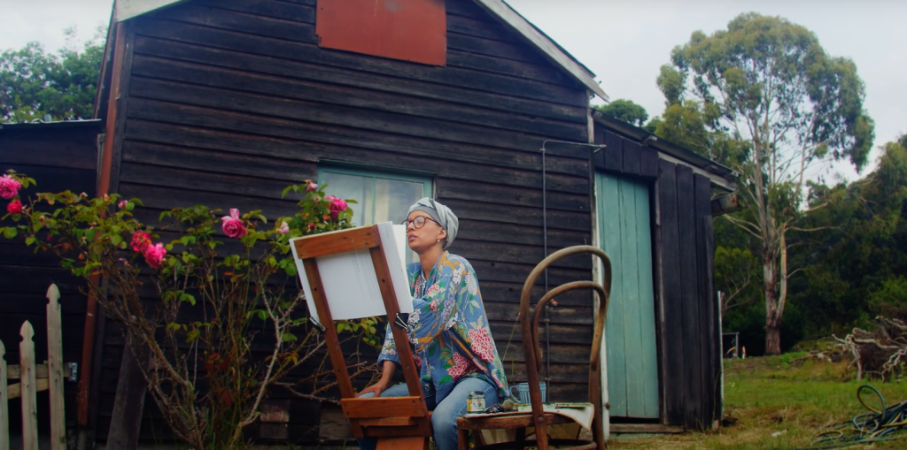 A person wearing glasses, a headscarf, and colorful clothes sits outdoors painting on an easel beside a rose bush, in front of a rustic wooden building with green doors. Trees are visible in the background.