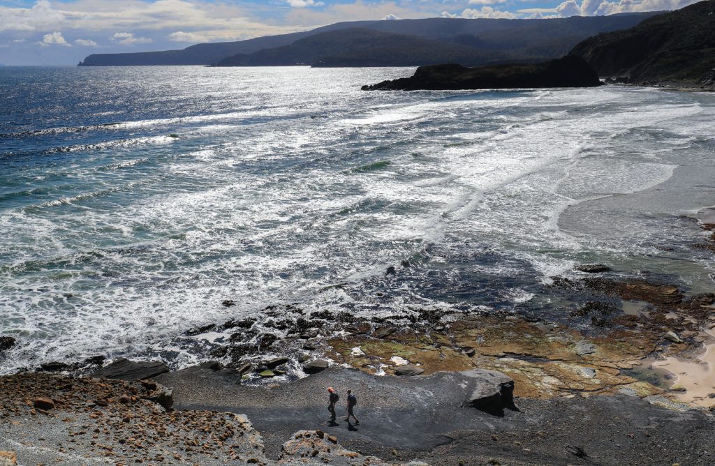 Two people walk along a rocky shoreline with waves crashing nearby, under a partly cloudy sky, with mountains and cliffs in the background.