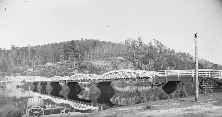 A black-and-white photo of a metal truss bridge crossing a river, with trees and hills in the background. The bridge and surroundings are reflected in the calm water below.