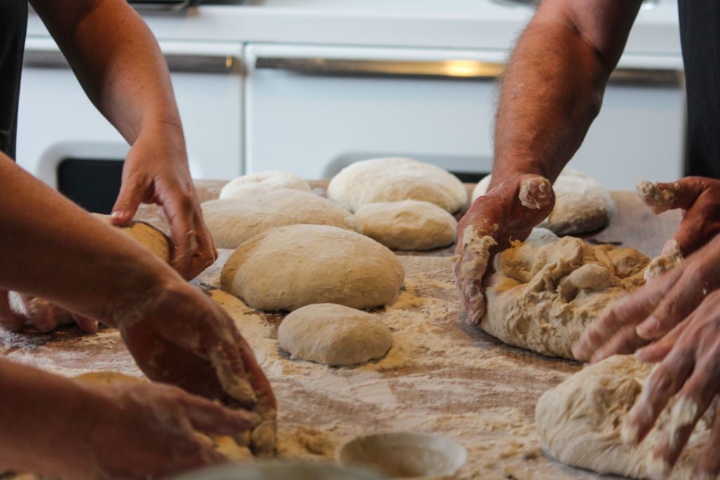 Three people knead and shape dough on a floured surface, with several dough balls and flour scattered around, suggesting bread or pizza preparation in a kitchen setting.