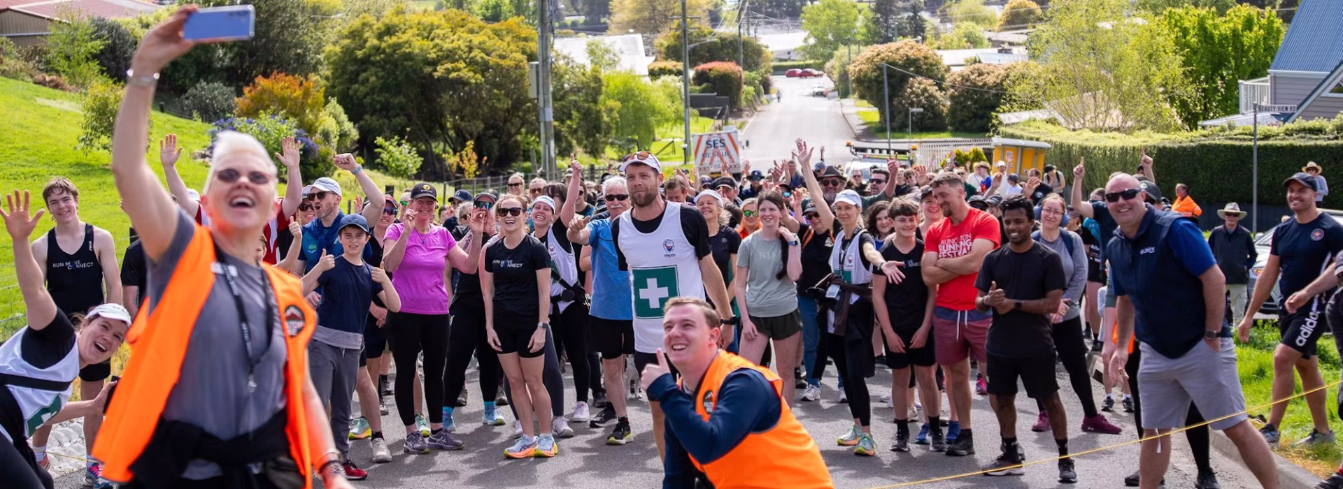 A group of people gathers outdoors on a sunny day, smiling and posing for a group selfie before a race or fun run. Two people in orange vests stand in front, while the crowd waves and looks at the camera.