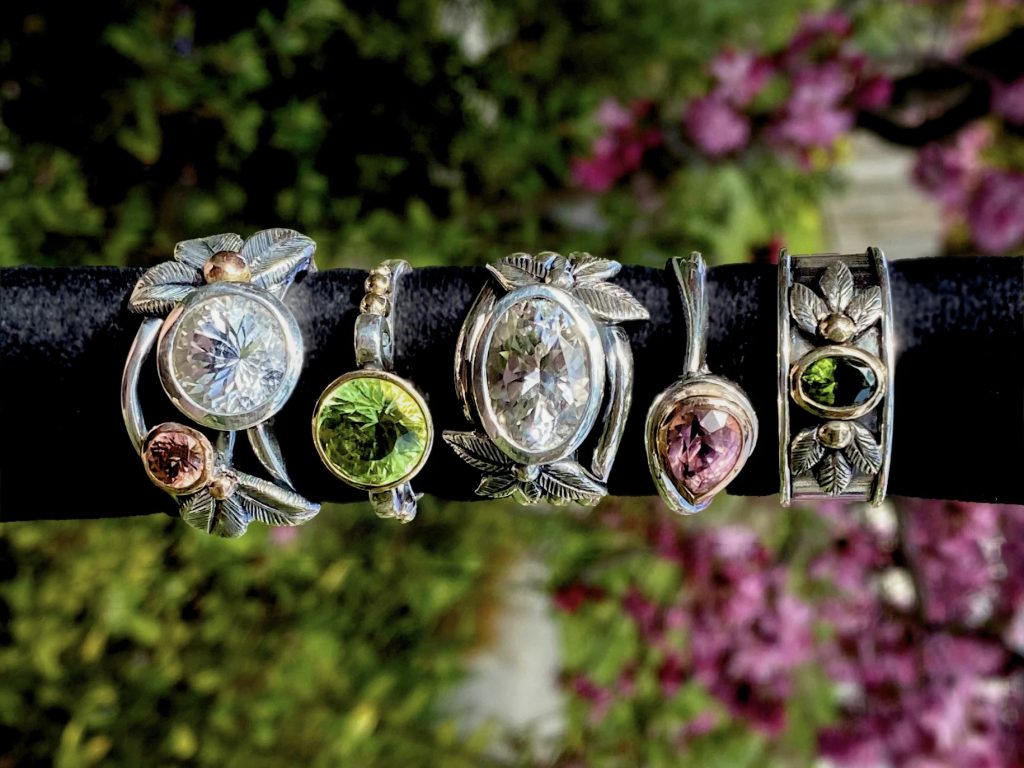 Five silver rings with colorful gemstones (clear, green, and pink) and leaf designs are displayed on a black bar. The background is blurred greenery and purple flowers.