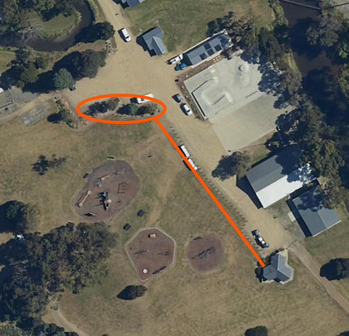 Aerial view of a park with playgrounds, nearby buildings, and trees. An orange line connects a small building to a circled area near a road and several parked vehicles.