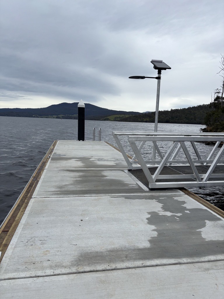 Concrete dock with metal railings extends over a calm lake under a cloudy sky. Hills and trees are visible in the background. A lamp post and a black piling are on the dock.