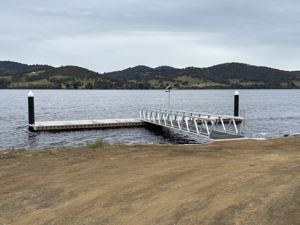 A floating dock with a metal ramp extends into a calm lake, surrounded by grassy hills under a cloudy sky. The shoreline is bare and unpaved.