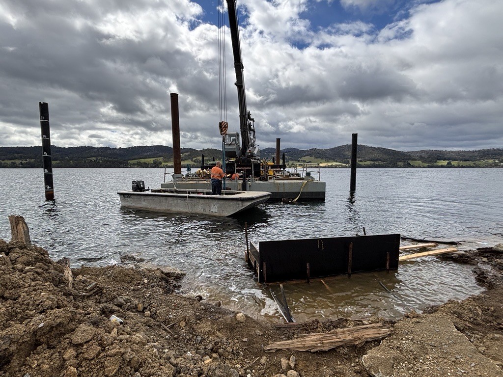 Workers operate a crane on a floating platform in a body of water, with construction equipment and materials nearby. The shoreline is visible in the foreground, and hills under a cloudy sky are in the background.