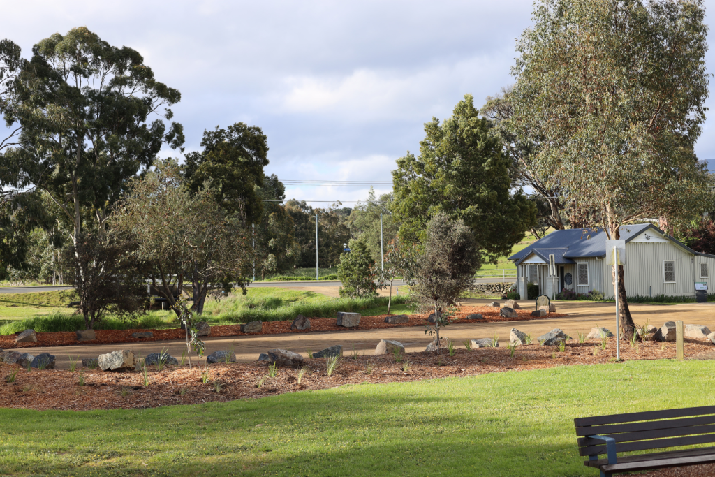 A small, light-colored house sits near a dirt driveway, surrounded by trees, rocks, and landscaping. A grassy area with a wooden bench is in the foreground under a partly cloudy sky.