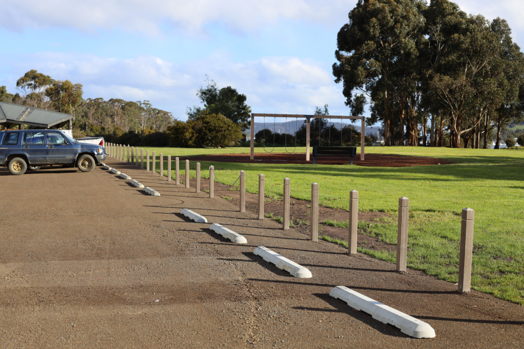 A row of parking spaces with concrete wheel stops lines a gravel lot next to a grassy park area with trees and a swing set in the background. A blue SUV is parked on the left.