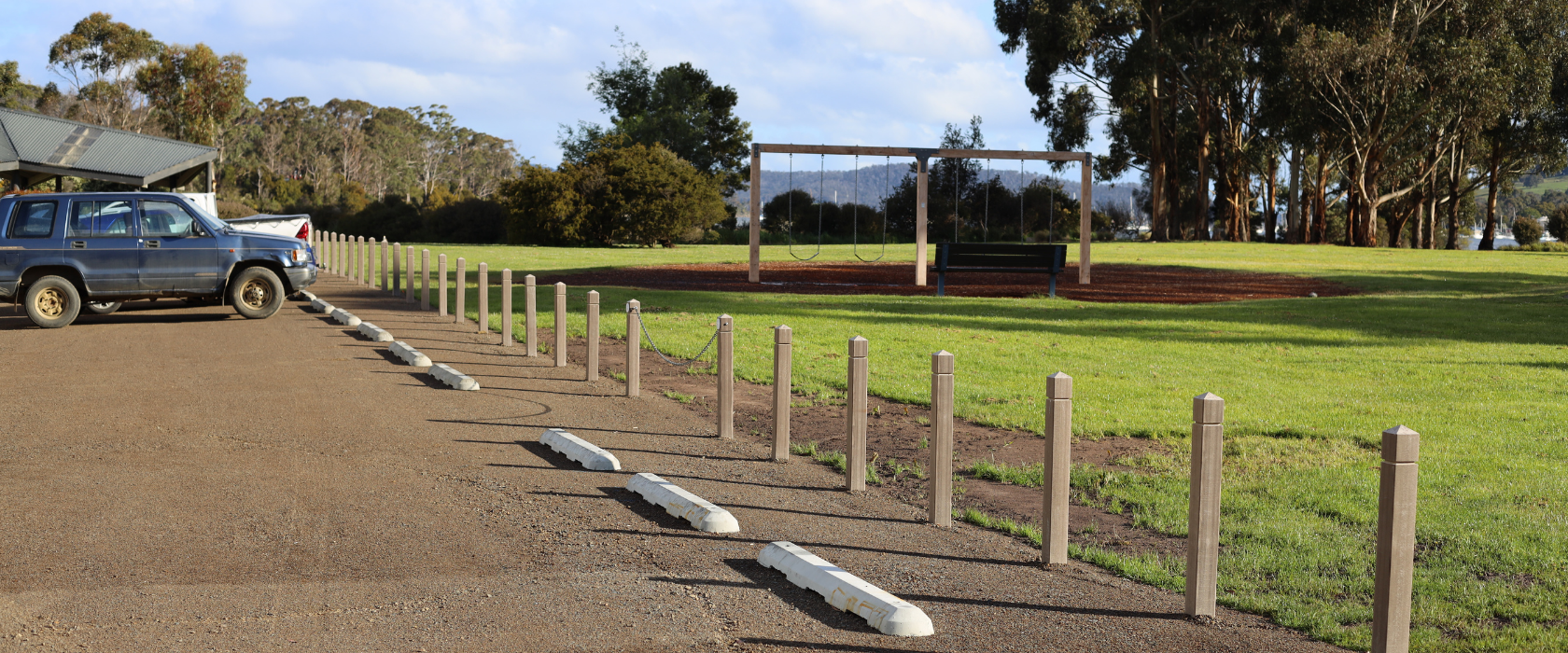 A row of parking spaces with concrete wheel stops lines a gravel lot next to a grassy park area with trees and a swing set in the background. A blue SUV is parked on the left.