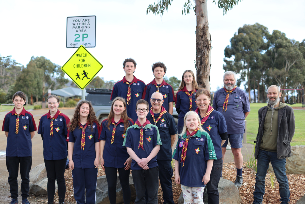 A group of scouts and adults in uniform pose outdoors on a footpath near rocks and grass. Behind them are street signs and trees. Everyone is smiling at the camera.