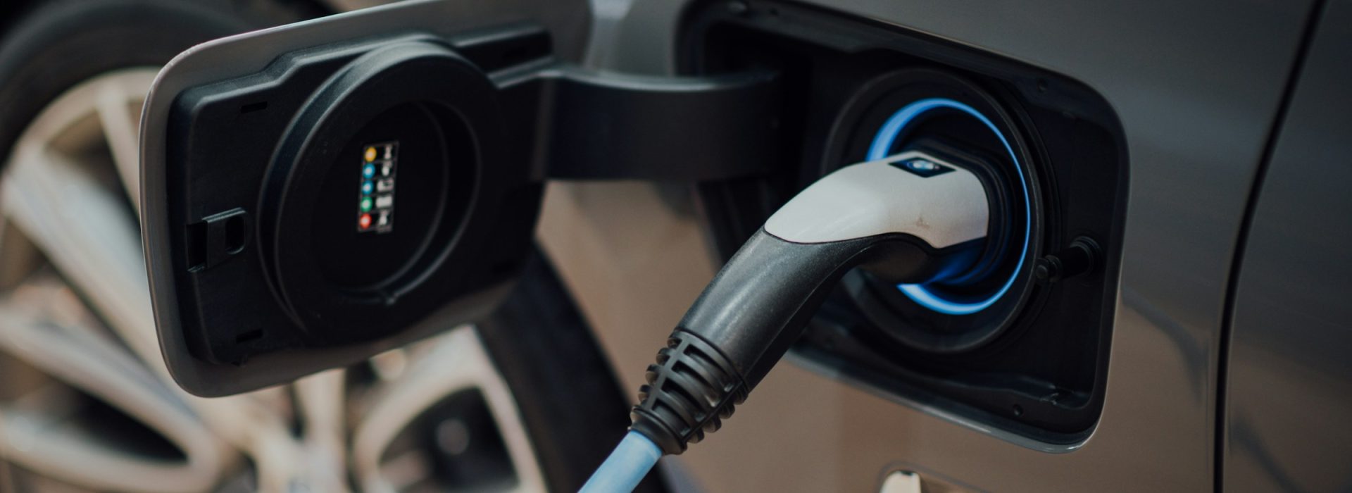 A close-up of an electric vehicle being charged, showing a charging cable plugged into the car’s charging port on a gray vehicle. The car’s wheel is partially visible in the background.