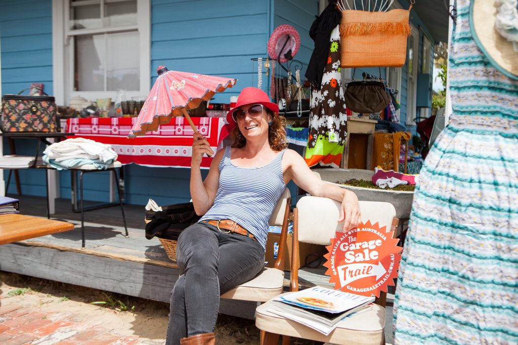 A smiling woman in a red hat sits on a chair holding a pink umbrella at a garage sale. She is surrounded by vintage items and a sign that reads “Garage Sale Trail” in front of a blue house.