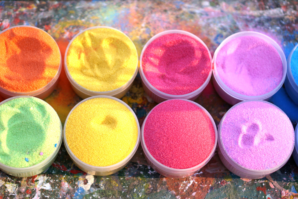 Eight circular containers filled with brightly colored sand—orange, yellow, pink, purple, green, and red—sit on a splattered, multicolored surface. Some sand containers show gentle imprints on their surfaces.